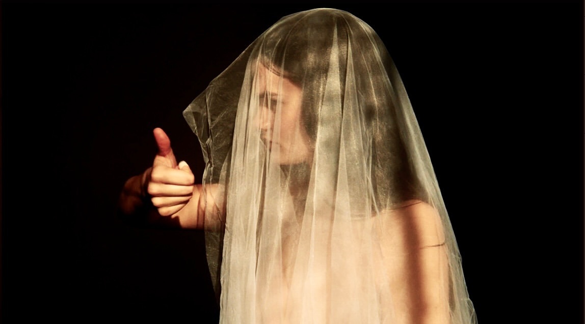 Dramatic portrait of a person with a sheer fabric veil giving a thumbs-up against a stark black background with light and shadow play.