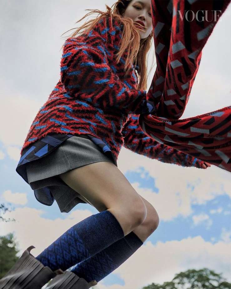 Young woman in mid-air wearing a patterned jacket, grey skirt, patterned socks, and boots, captured in an outdoor setting with Vogue logo.