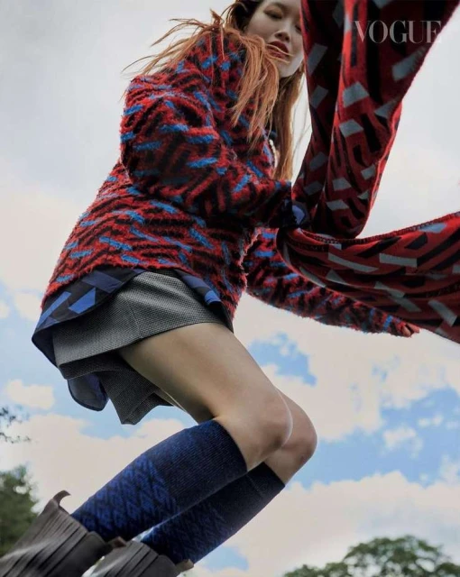 Young woman in mid-air wearing a patterned jacket, grey skirt, patterned socks, and boots, captured in an outdoor setting with Vogue logo.