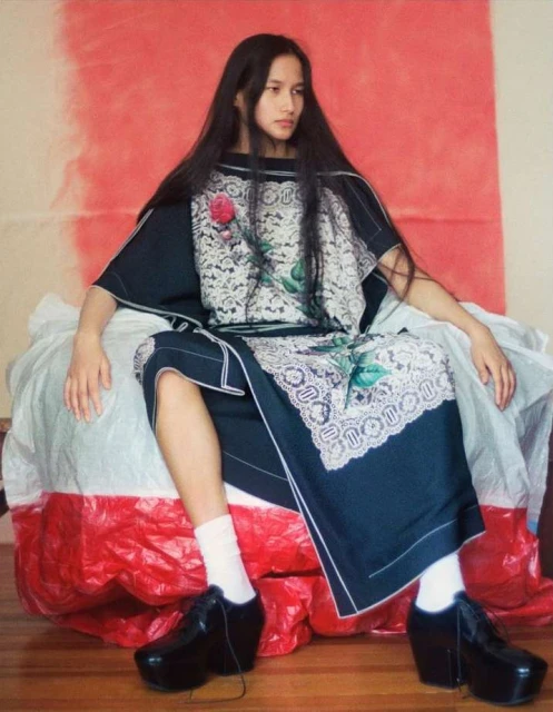 A woman with long black hair sits in a unique dress and platform shoes against a backdrop of red and cream colors in an indoor setting.