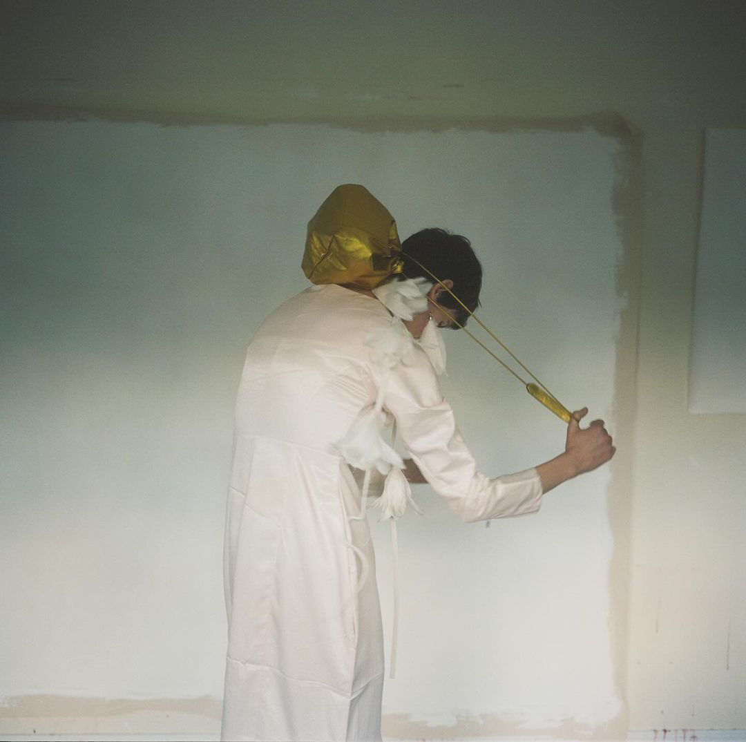 Person in lab coat prepares to launch golden balloon with slingshot in bright room, artistic studio shot, unique and slightly surreal aesthetic.