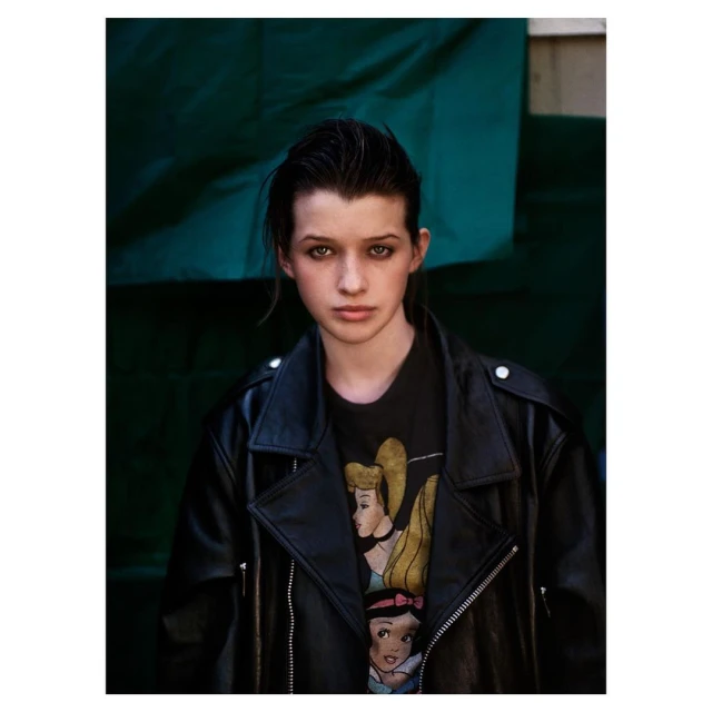 Portrait of a young woman with dark hair and a leather jacket, featuring a t-shirt with cartoon princess graphics against a green backdrop.