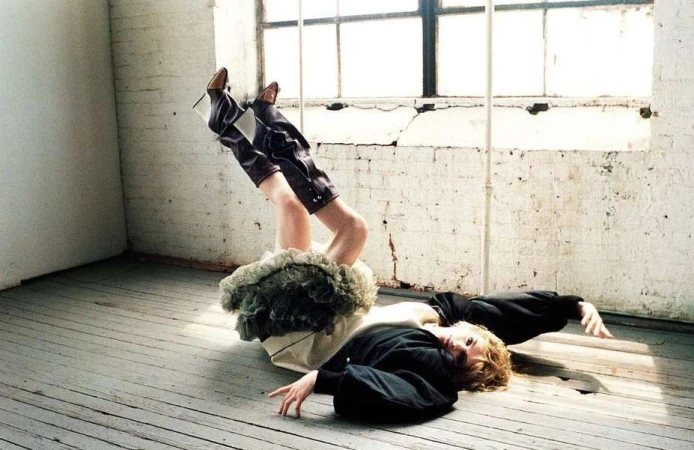 Model lying on wooden floor with legs raised in the air, wearing dark boots and a ruffled skirt.
