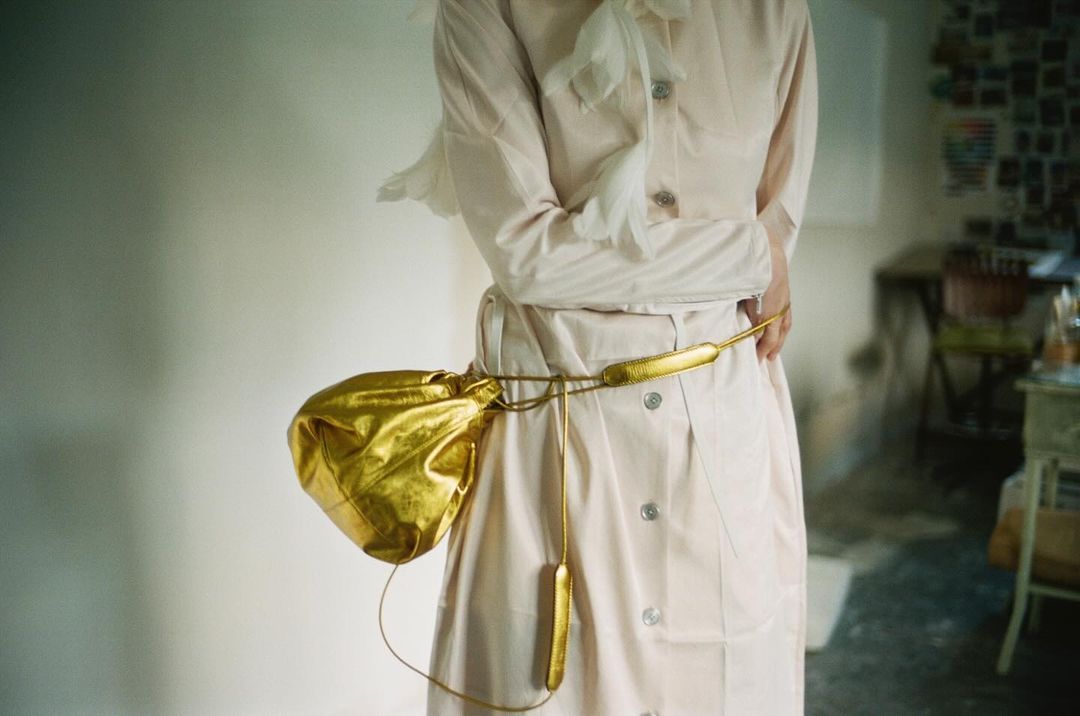 A woman is wearing a light pink dress with a gold belt and a gold purse in an indoor setting with soft, natural lighting.