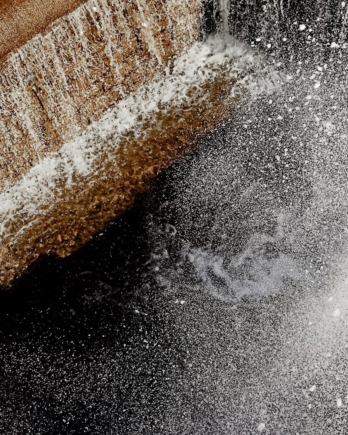 Waterfall flowing over a concrete wall with a dramatic display of water droplets and spray, against a dark background.