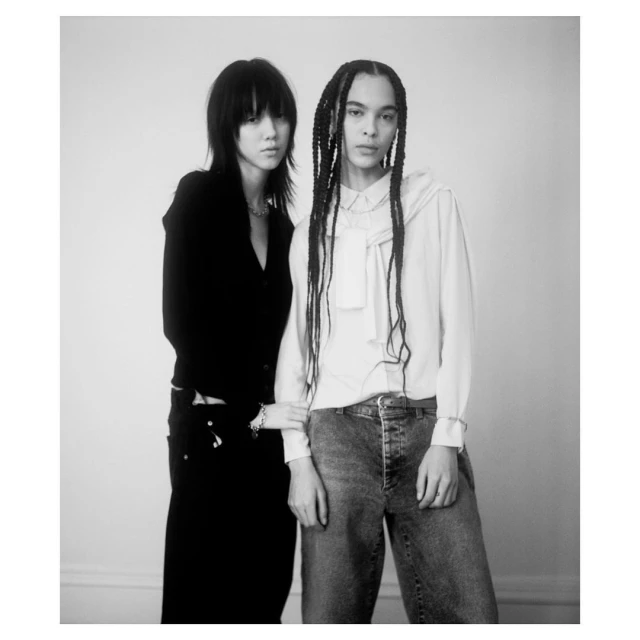 Black-and-white studio shot of two young adults in casual fashion: one in black top, jeans, and the other in jeans and a white blouse.