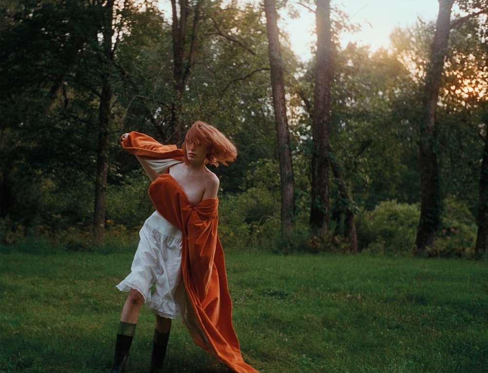 An adult woman in a white dress and orange shawl stands in a grassy field in front of a forest during golden hour.