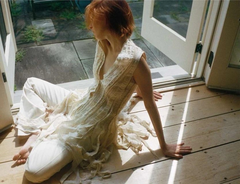 A fair-skinned woman with red hair poses indoors near open doors wearing a white dress and white pants, with sunlight on the wood floor.