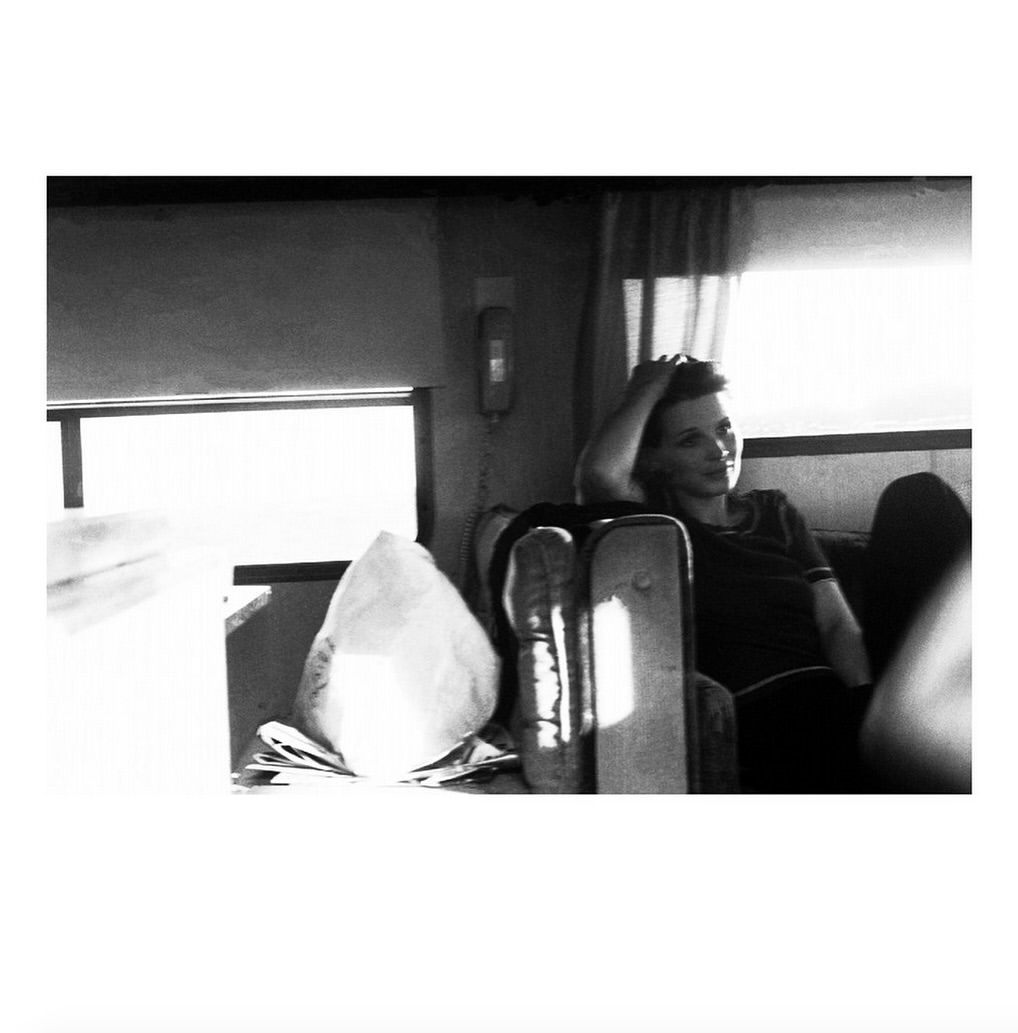 Monochrome shot of a woman seated indoors in casual attire, looking off-camera with her hand on her head, alongside luggage.