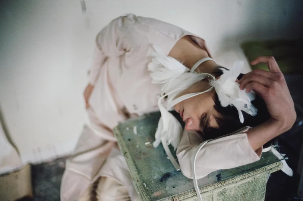 Artistic image of a person lying on a green textured surface, head wrapped in white fabric with feathers, pale tones, and soft focus.
