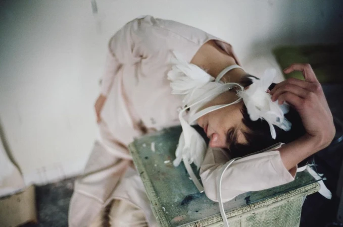 Artistic image of a person lying on a green textured surface, head wrapped in white fabric with feathers, pale tones, and soft focus.