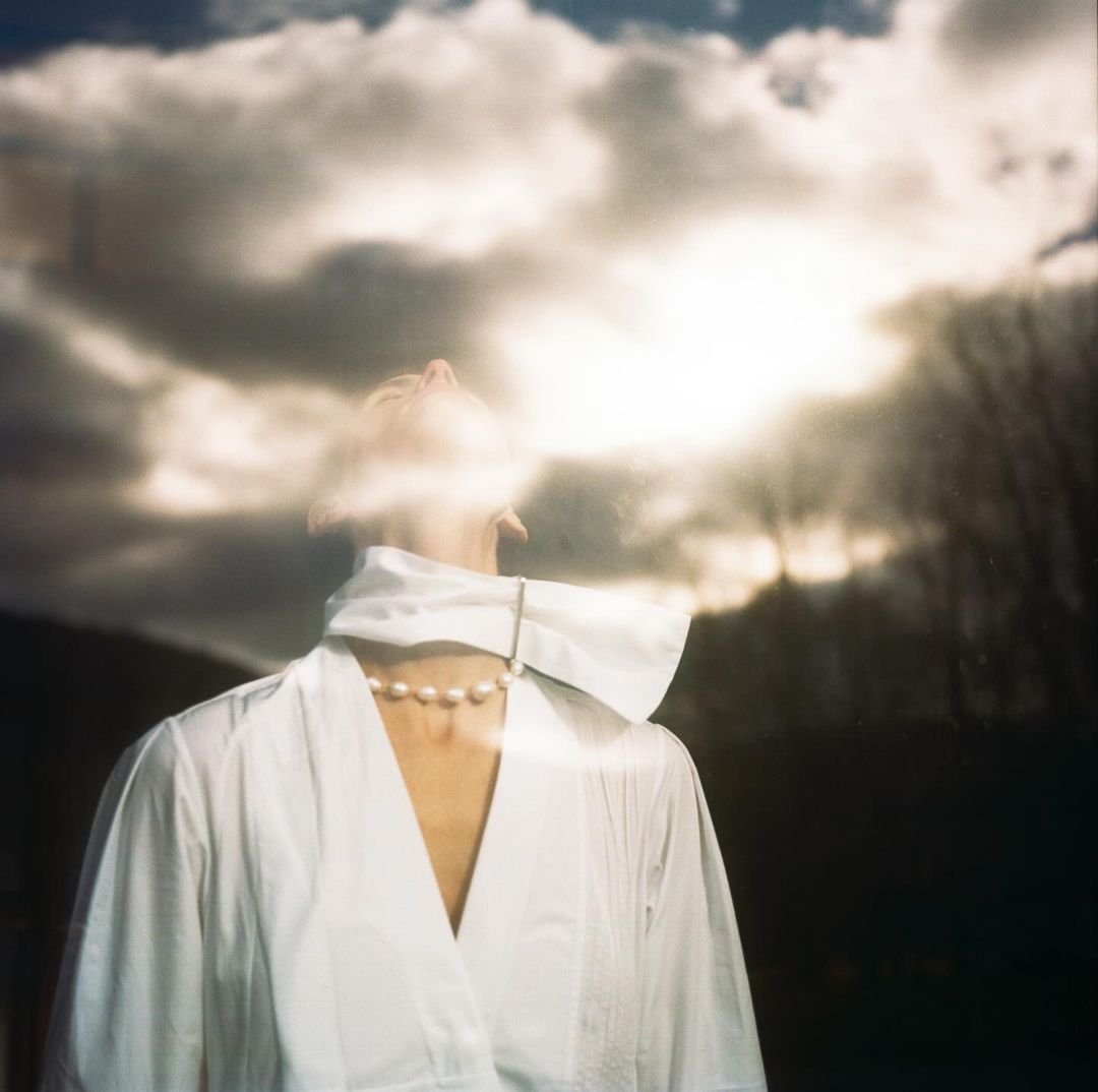 Adult female with cloudy head facing upward, wearing white clothing and pearl necklace, standing outdoors with blurred natural background.