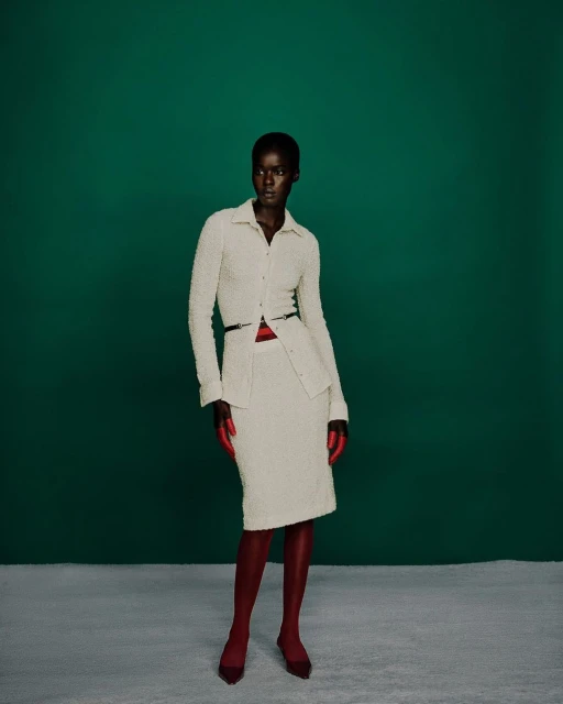A dark-skinned woman poses in a cream-colored skirt suit with a matching blouse, a thin black belt, and reddish-brown tights and shoes.