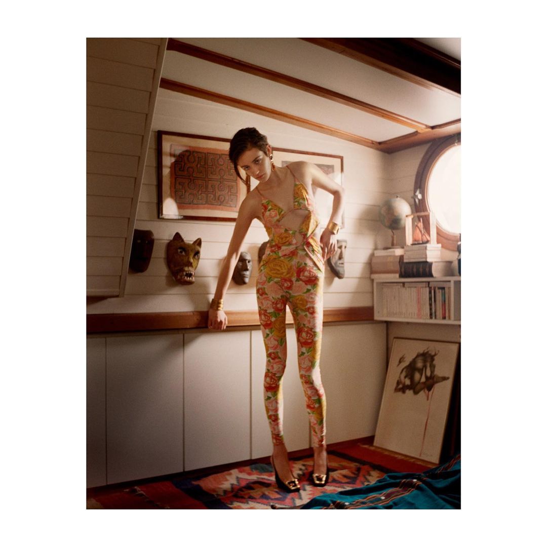 A young woman poses in a floral jumpsuit and heels in a stylish indoor setting with masks and artwork; fashion editorial with natural light.