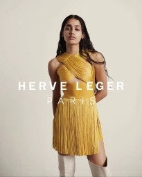 Model in a yellow fringed minidress and white boots standing against a neutral background by Drew Jarrett