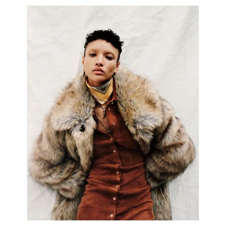 Model wearing a faux fur collar coat and rust button-up dress with a patterned neck scarf against a textured white background.