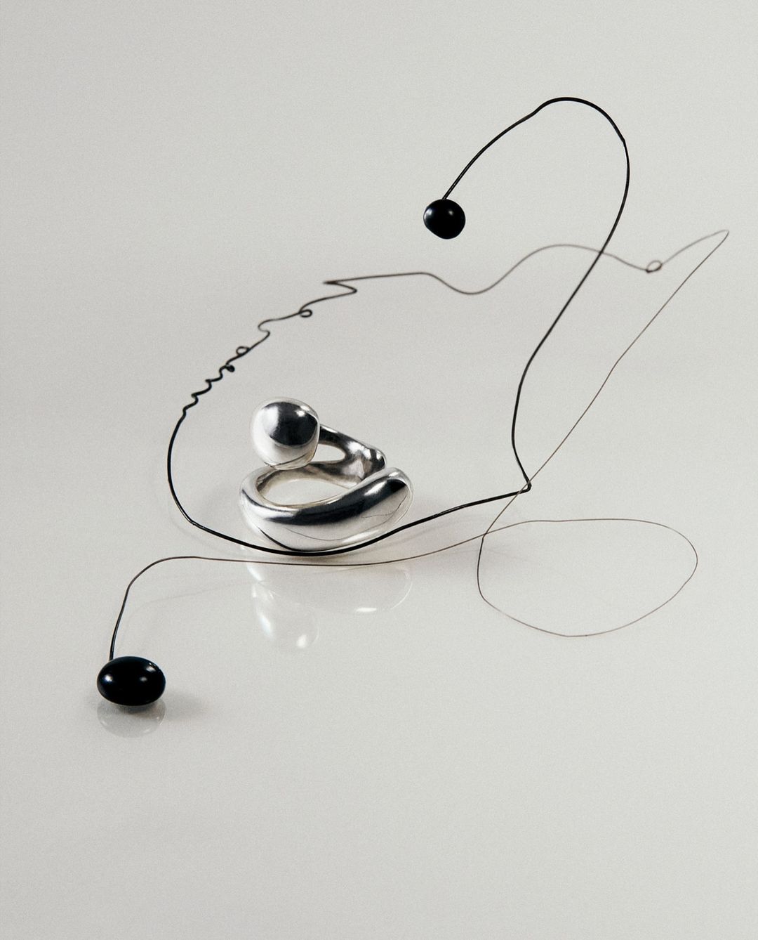 Abstract jewelry product shot: a silver ring with a sphere detail rests on a white surface accented by a thin black wire.