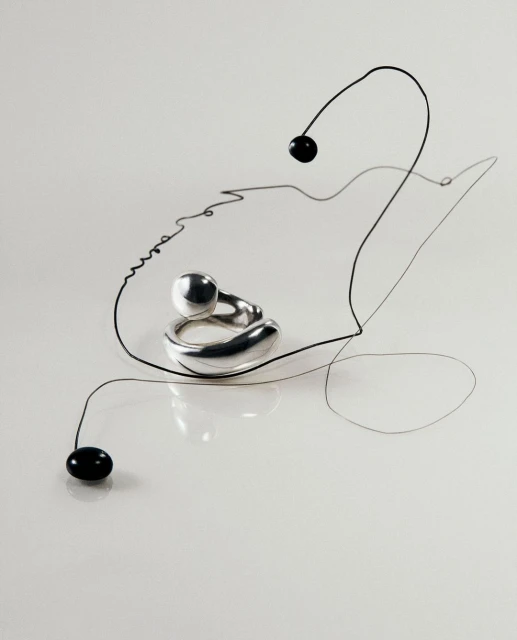 Abstract jewelry product shot: a silver ring with a sphere detail rests on a white surface accented by a thin black wire.
