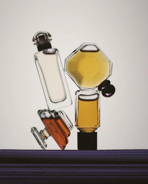 Four perfume bottles of different shapes and colors arranged on a textured purple surface against a gradient white background in a studio setting.