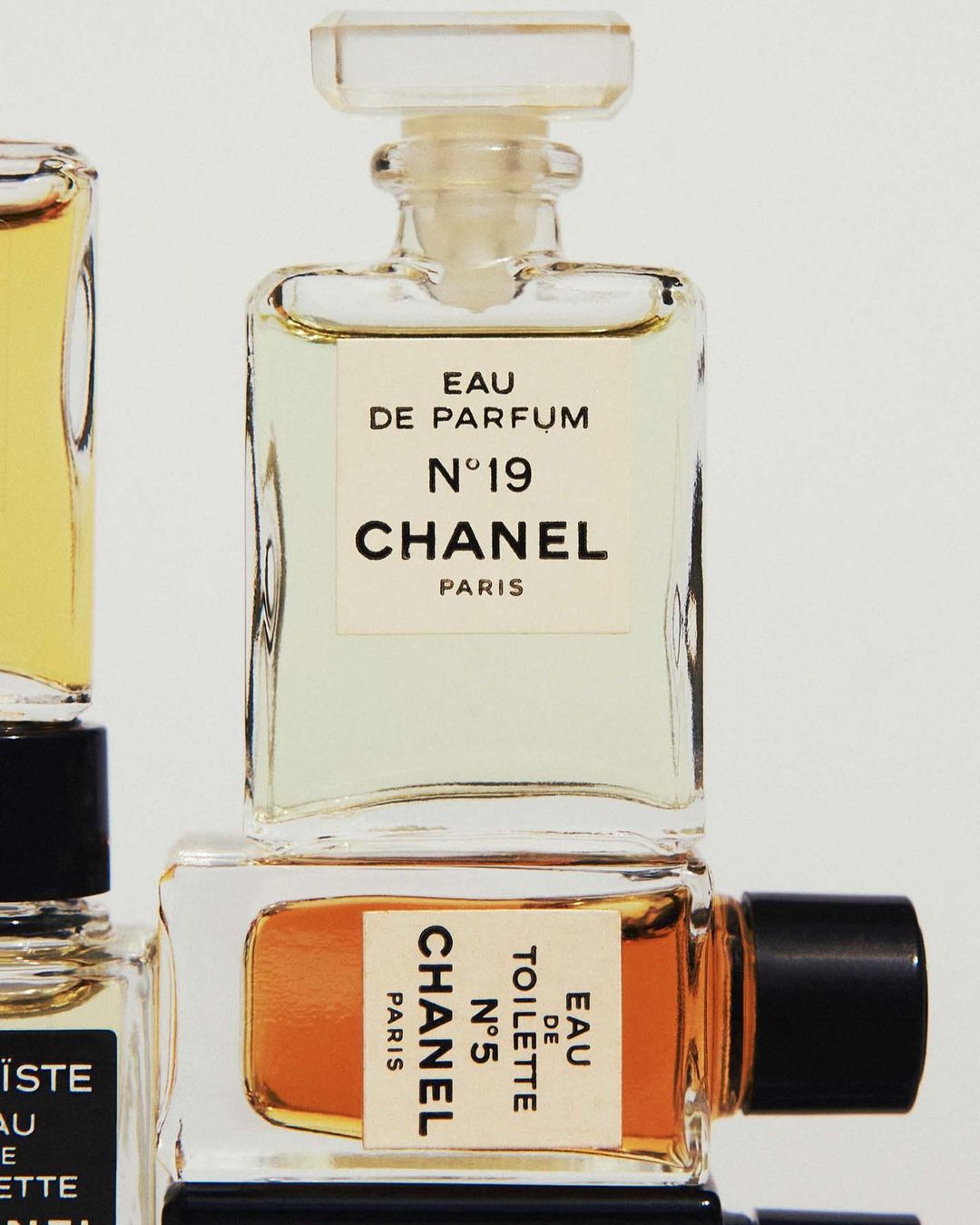 Close-up studio shot of Chanel N°5 and N°19 Eau de Parfum bottles with clear glass, white labels, and golden and amber liquid tones.