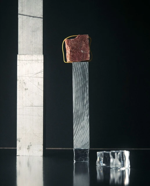 Abstract still life photography of a red rock secured with a gold wire on a textured glass post with a black background.