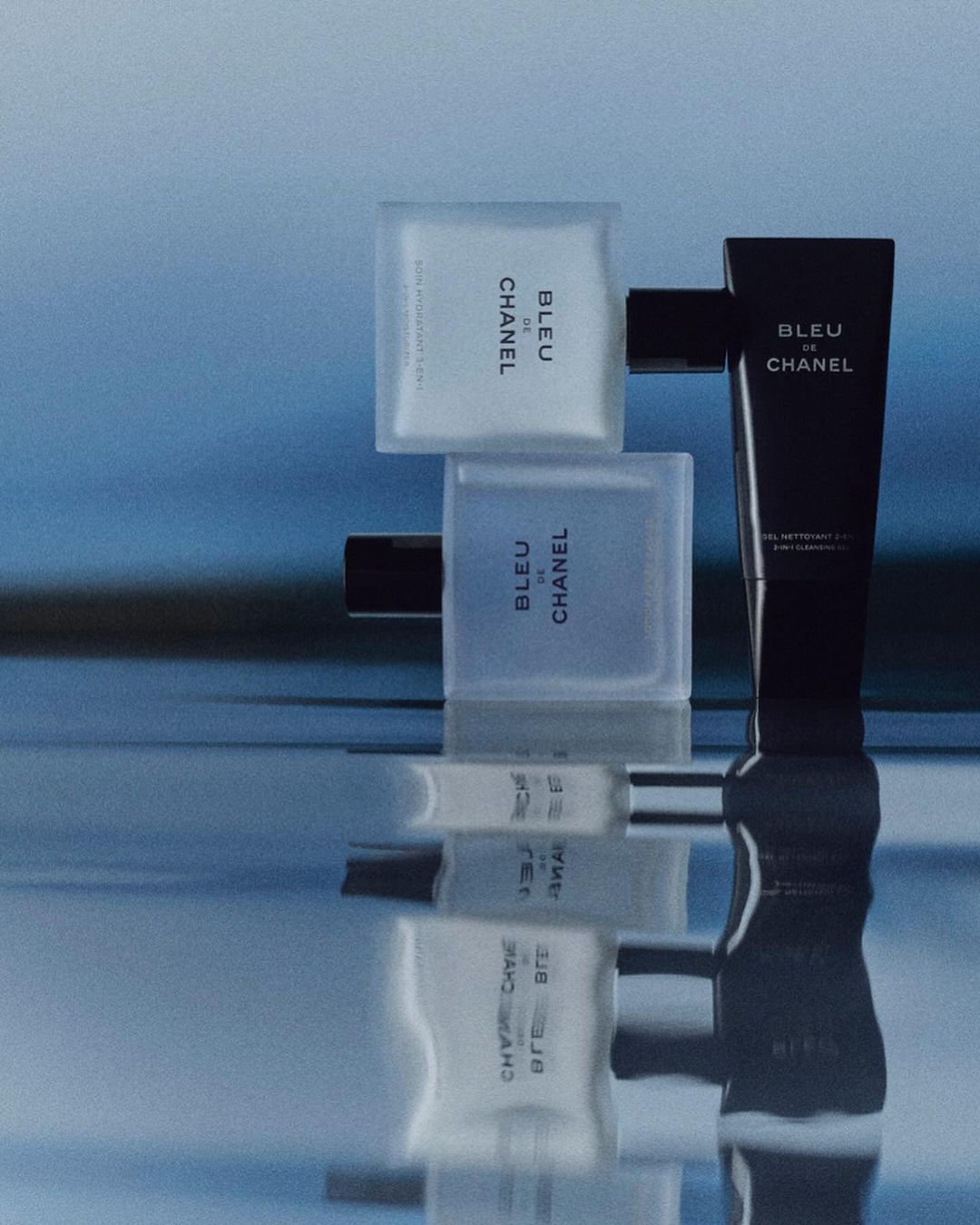 Bleu de Chanel product presentation on reflective surface. Includes perfume bottle, body wash and aftershave lotion against blue background.