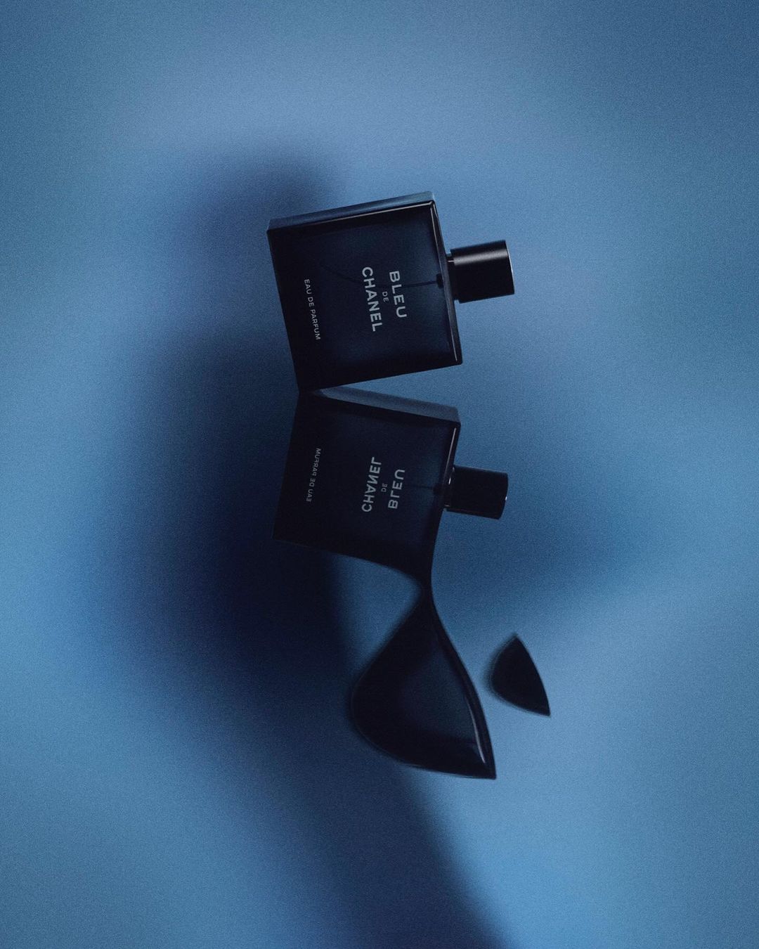 Two Chanel Bleu de Chanel bottles, one upright and one reflected, on a bright blue surface, showcasing a men's fragrance product shot.