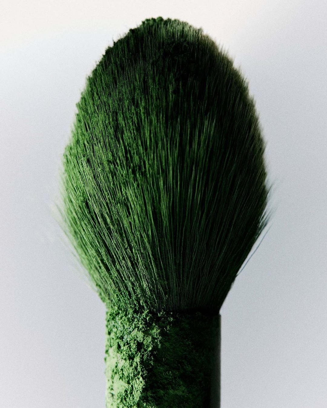 Close-up of a green makeup brush coated in vibrant green powder, set against a neutral background in a beauty context.