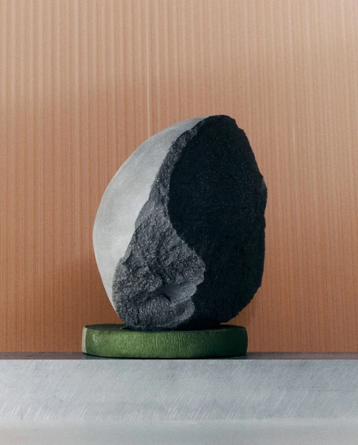 A stone sculpture sits atop a textured green stand in a modern composition, complemented by a neutral, vertically-lined background.