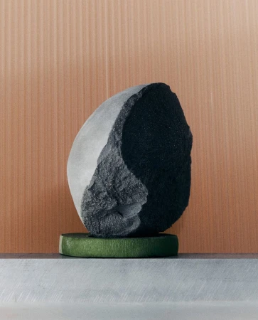 A stone sculpture sits atop a textured green stand in a modern composition, complemented by a neutral, vertically-lined background.