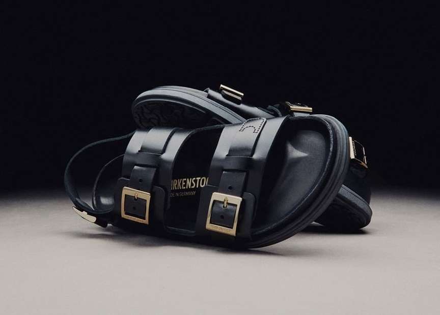 Close-up of black leather Birkenstock sandals with gold buckles on a neutral surface against a dark background. Studio product shot.