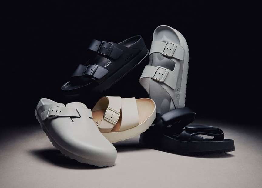 A product shot featuring an assortment of casual sandals in black, grey, and beige arranged on a neutral surface against a dark backdrop.
