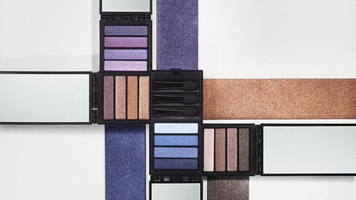 Flat lay of four open eye shadow palettes with various colors arranged in a square shape on a solid off-white surface with glitter accents.