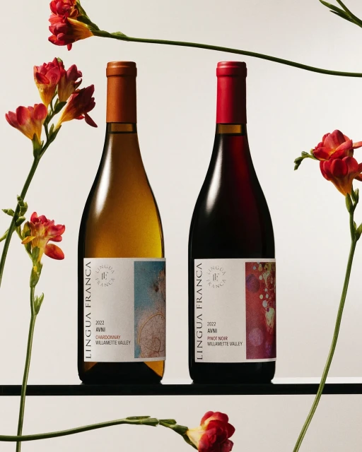 Two bottles of Lingua Franca wine on a black shelf surrounded by red flowers against a neutral background in a product studio shot.