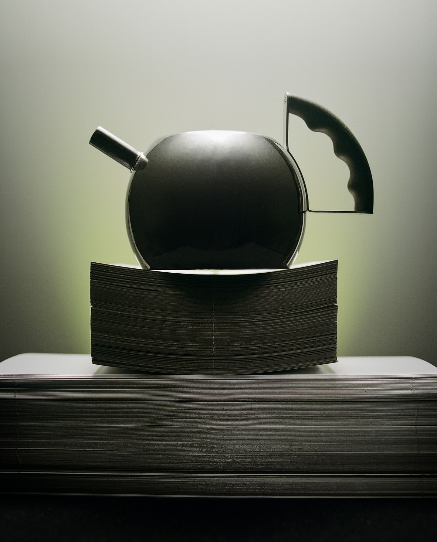 Studio shot of a black teapot on a pile of paper with soft lighting, creating a high-contrast image emphasizing form and texture.