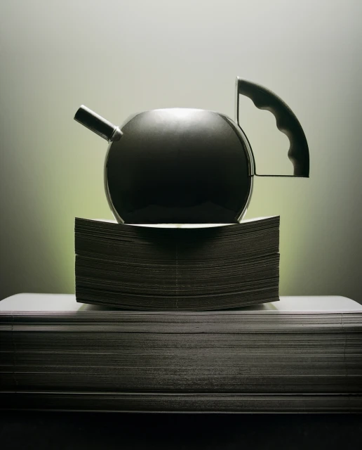 Studio shot of a black teapot on a pile of paper with soft lighting, creating a high-contrast image emphasizing form and texture.