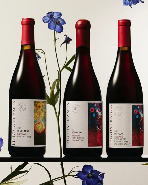 Three bottles of Lingua Franca Pinot Noir wine with red capsules, arranged alongside delicate blue Larkspur flowers in a studio setting.