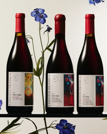 Three bottles of Lingua Franca Pinot Noir wine with red capsules, arranged alongside delicate blue Larkspur flowers in a studio setting.