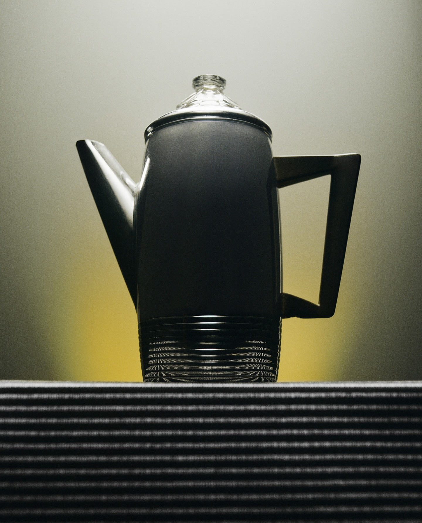 Elevated view of a grey coffee pot with a clear lid sitting on a striped surface against a gradient yellow background.