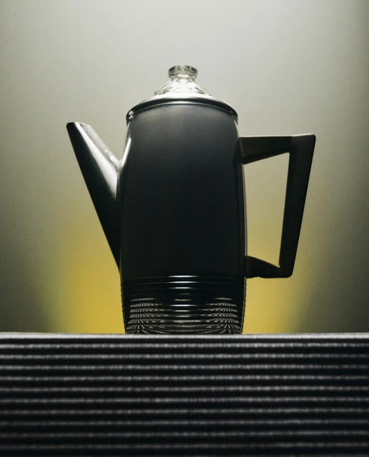 Elevated view of a grey coffee pot with a clear lid sitting on a striped surface against a gradient yellow background. 