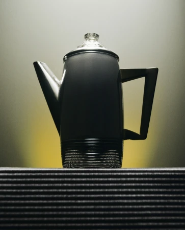 Elevated view of a grey coffee pot with a clear lid sitting on a striped surface against a gradient yellow background. 