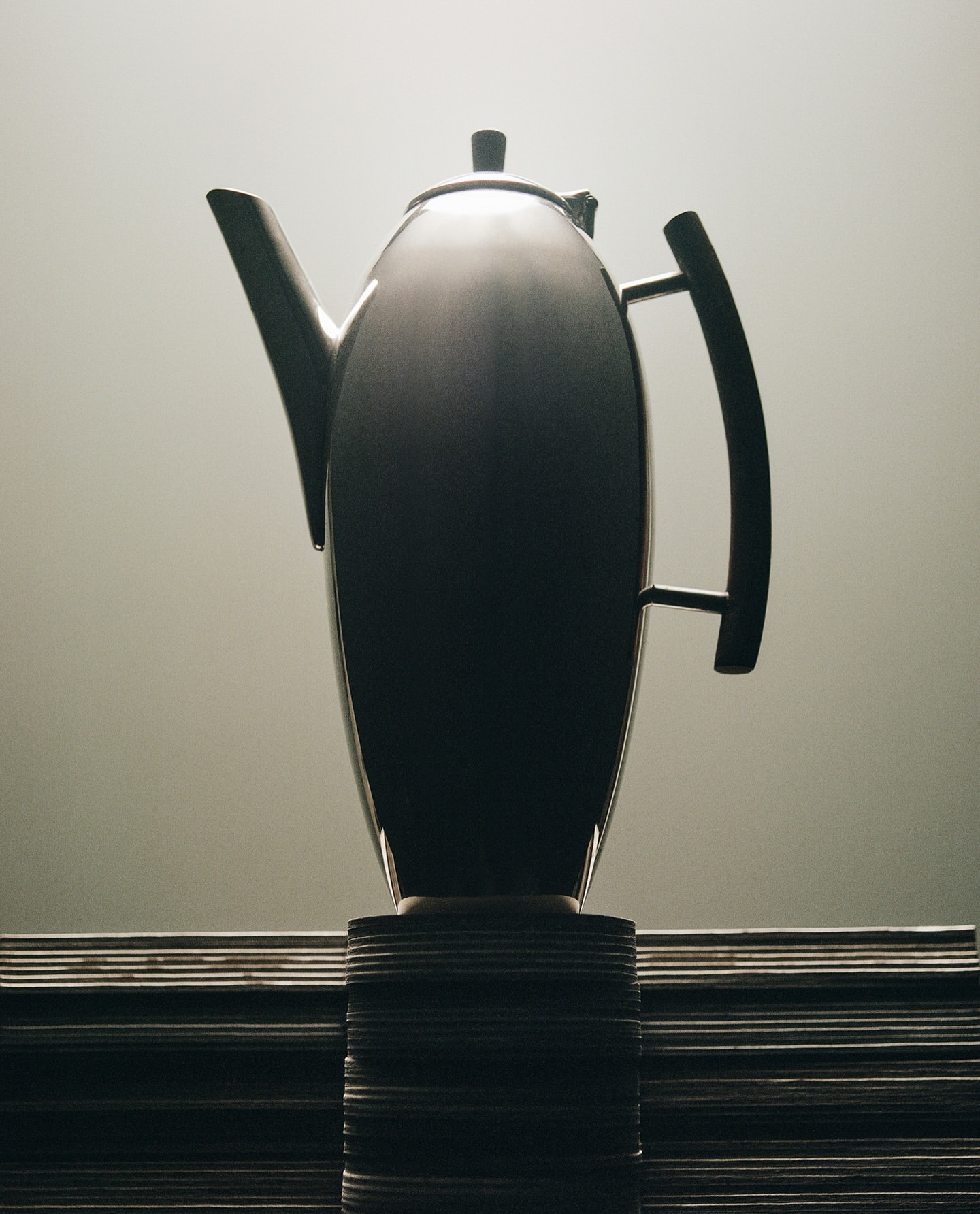 Elegant silver teapot with curved handle and spout, displayed on a stack of plates against a soft gradient gray background.