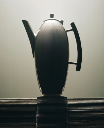 Elegant silver teapot with curved handle and spout, displayed on a stack of plates against a soft gradient gray background.