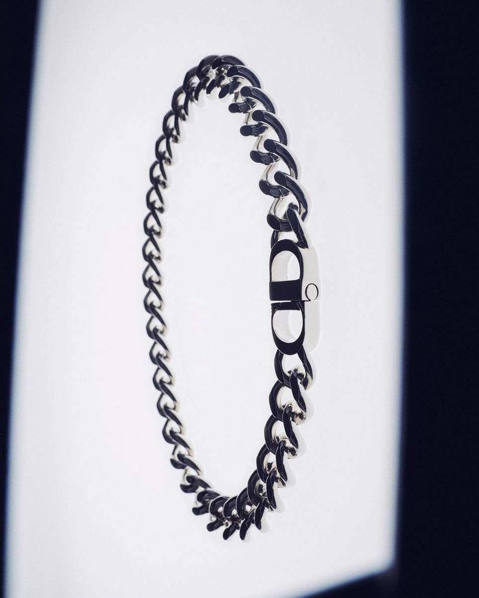 Close-up of a silver chain necklace with a silver CD logo charm on a white background with black gradient lighting.