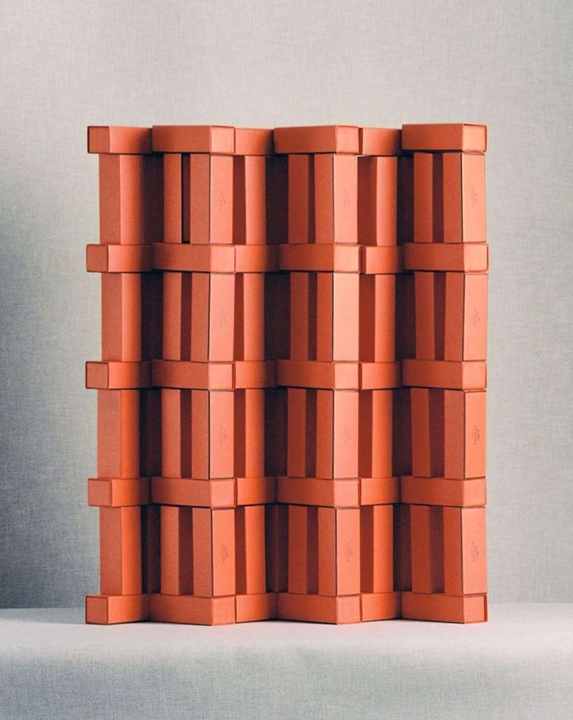 Stacked orange boxes create a geometric, three-dimensional wall, resembling an art installation with a textured, paper-like quality in soft lighting.