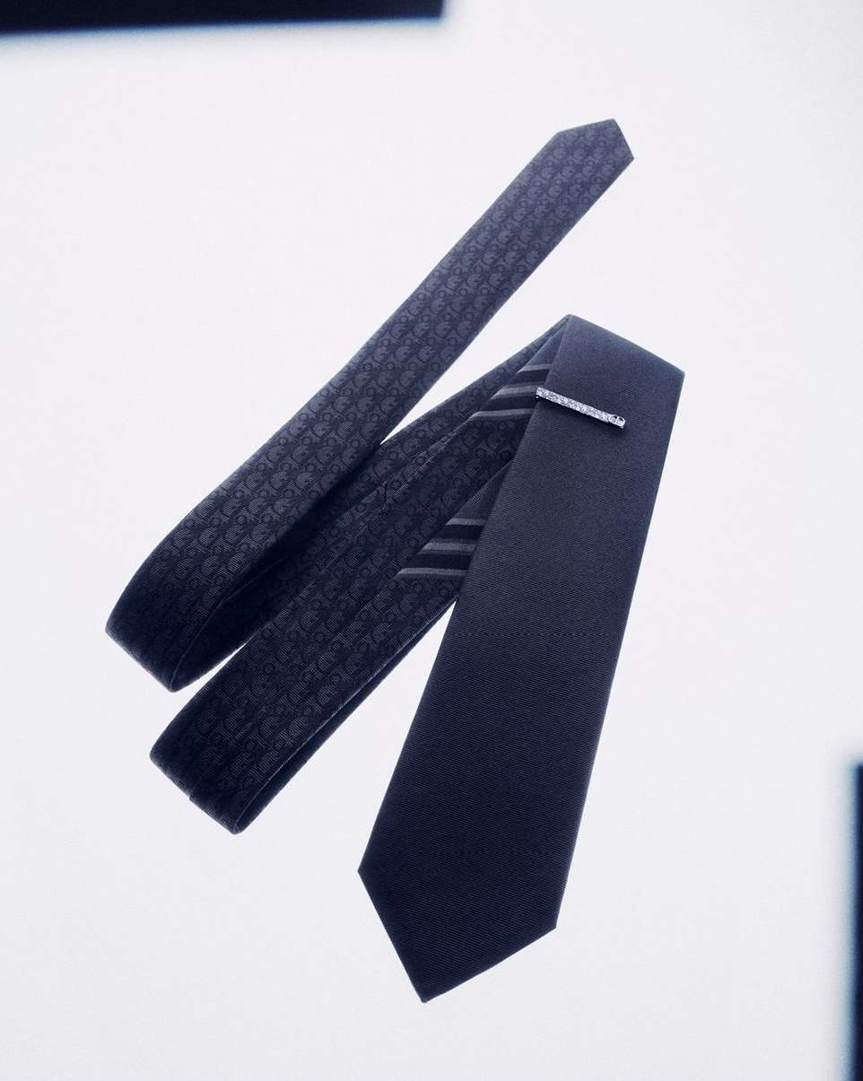 Two dark blue silk ties, one patterned, the other with a tie clip, rest on a white surface, casting subtle shadows.