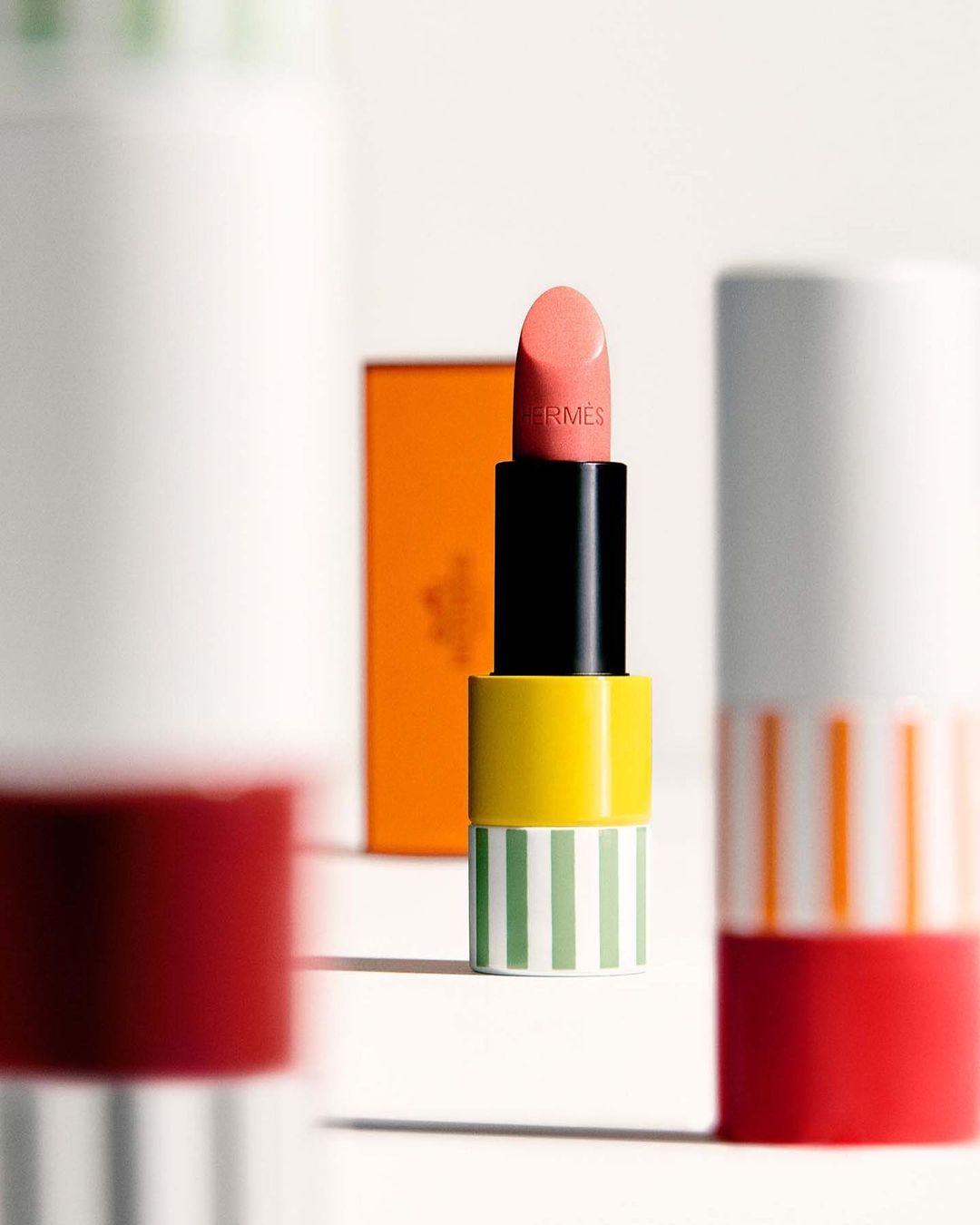 Hermès lipstick tube in peach, yellow, green, and black on a white surface with other lipstick tubes in the background.