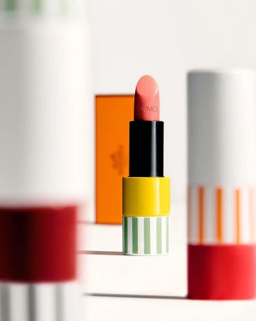 Hermès lipstick tube in peach, yellow, green, and black on a white surface with other lipstick tubes in the background.