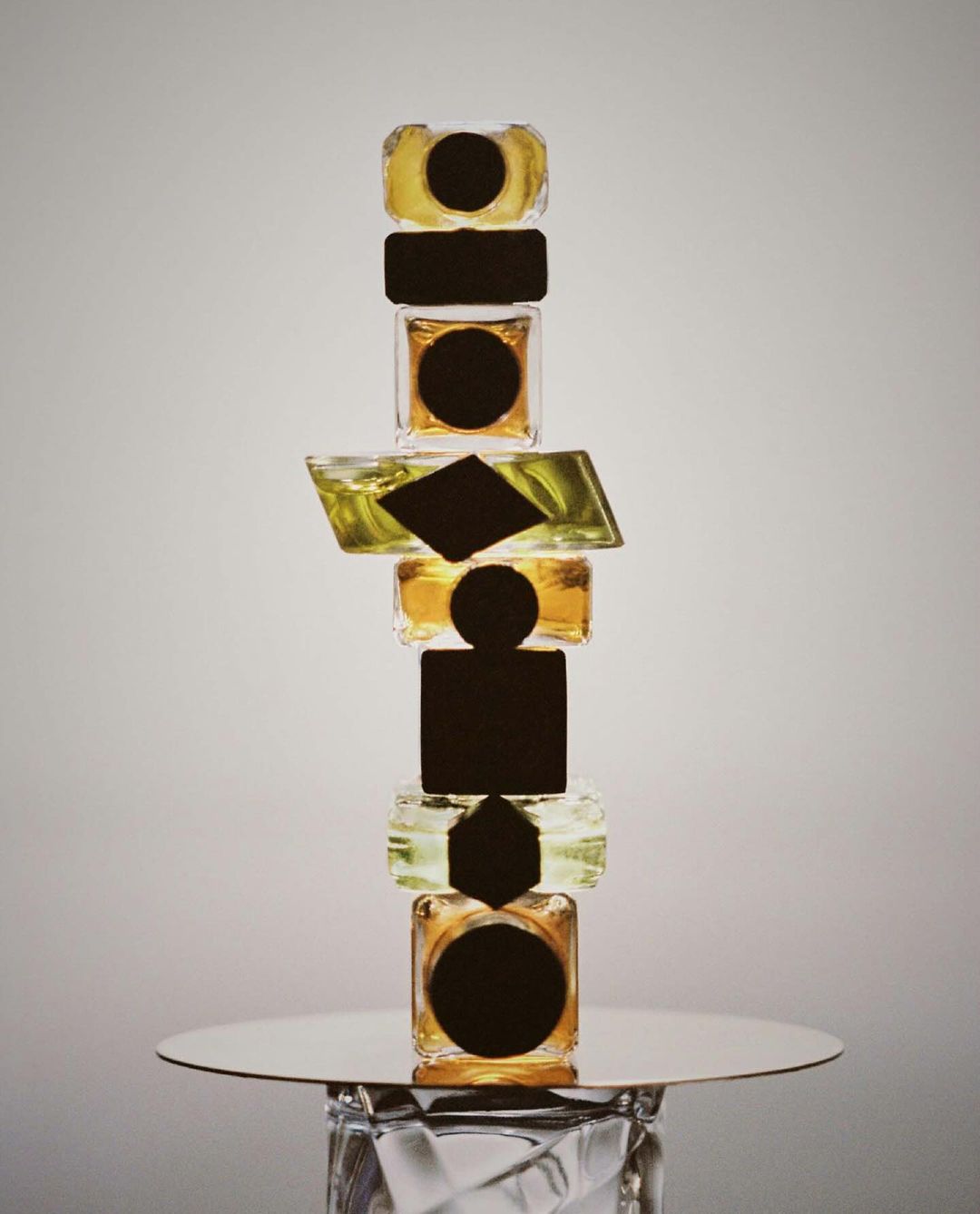 Tower of geometric-shaped glass jars on a reflective surface, featuring clear and yellow liquids and abstract black labels.