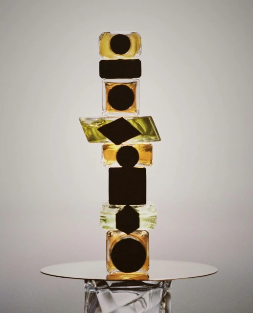 Tower of geometric-shaped glass jars on a reflective surface, featuring clear and yellow liquids and abstract black labels.